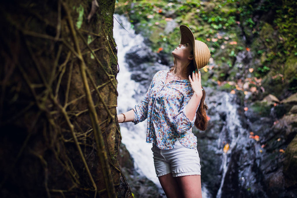 Beautiful young woman standing at the lake near waterfall in white dress and straw hat - Photo, Image