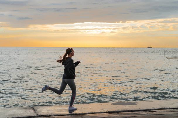 confident sports girl jogging on the seafront at sunset, listening to music on headphones - Фото, изображение