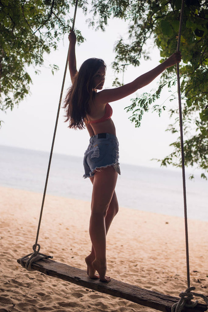 Nuori nainen seisoo köydellä keinu trooppinen hiekkaranta taustalla palmuja, sininen taivas ja meri, näkymä takaa
 - Valokuva, kuva