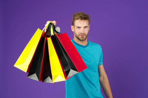 Shopping or sale and cyber monday. Man with shopping bags on violet background. Macho with colorful paper bags. Fashion shopper in casual blue tshirt. Holidays preparation and celebration. - Fotografie, Obrázek