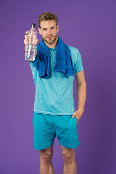 Man hold water bottle on violet background. Sportsman with plastic bottle. Athlete in blue tshirt and shorts with towel after training. Thirst and dehydration. Sport activity and energy. - Foto, immagini