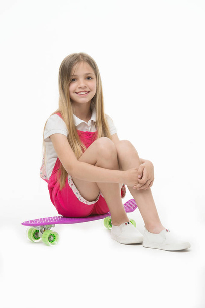 Small girl sit on skate board isolated on white. Child skater smile with longboard. Skateboard kid in pink jumpsuit. Sport activity and energy. Childhood and active games. Beauty on skateboard - Photo, Image