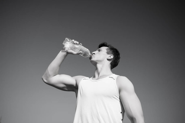 Athlete in green tshirt on blue sky. Sport and fitness concept. Bodybuilder with muscular hands muscles biceps, triceps. Healthy lifestyle and health. Man drinking water from bottle on sunny day. - Fotografie, Obrázek