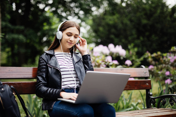 Student sitting on bench listening to music and using laptop and wearing headphones - 写真・画像
