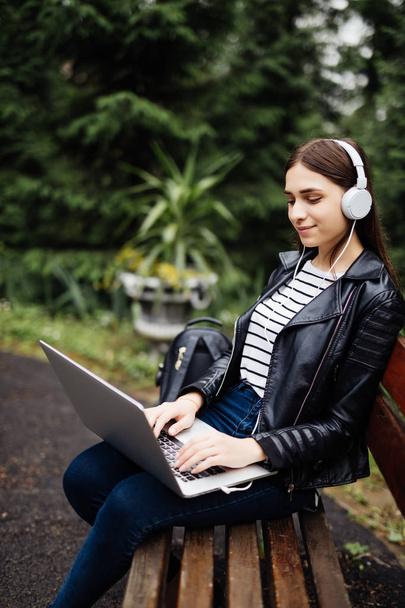 Estudiante sonriente sentado en el banco escuchando música y sosteniendo la computadora portátil en el parque en la escuela
 - Foto, imagen