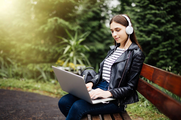 Mosolygó diák ül a padon, zene és gazdaság laptop, parkban, iskolában - Fotó, kép