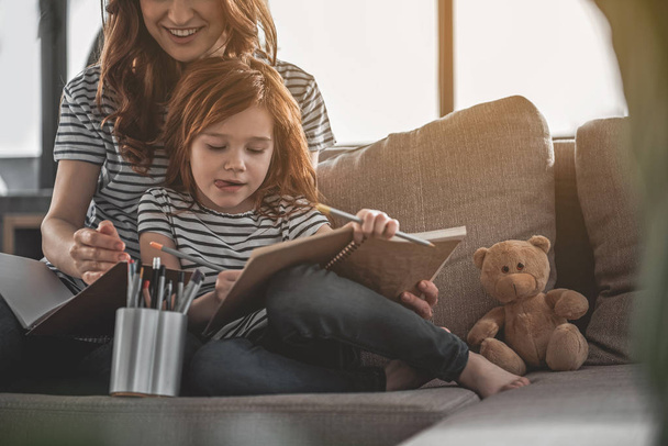 Small red-haired girl drawing intently with her mom nearby - Photo, Image