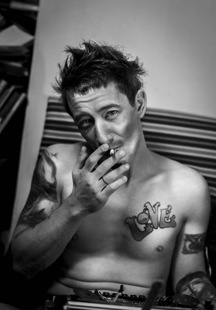 Verticale authentiek artistieke zwart-wit portret van knappe Kaukasische mannelijk model met sigaret in de hand close-up en roken om heen. Ondergrondse stijl man met tatoeage op het lichaam poseren binnenshuis. - Foto, afbeelding