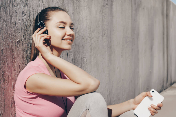 happy young girl resting after a hard workout outdoors, listening to music on headphones and smiling - Photo, Image