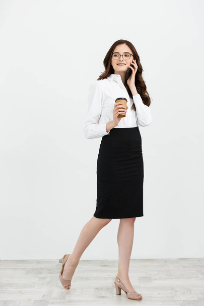 Full length Portrait of beautiful young business woman in formal wear walking and talking for cell phone with takeaway coffee in hand isolated over gray background - Фото, изображение
