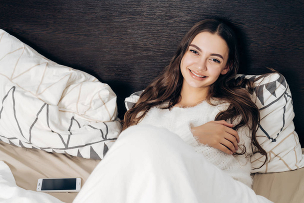 beautiful cute girl in white pajamas lies in bed, resting after a hard day's work and smiling - Fotografie, Obrázek