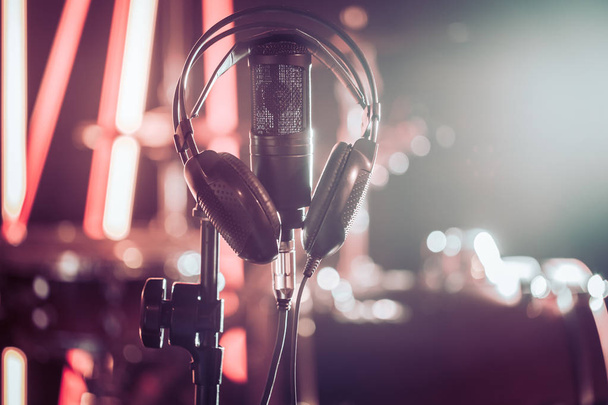 Studiomikrofon und Kopfhörer auf einem Nahaufnahme-Stativ, in einem Aufnahmestudio oder Konzertsaal, mit einem Schlagzeug im Hintergrund im Unschärfemodus. schöner verschwommener Hintergrund farbiger Lichter - Foto, Bild
