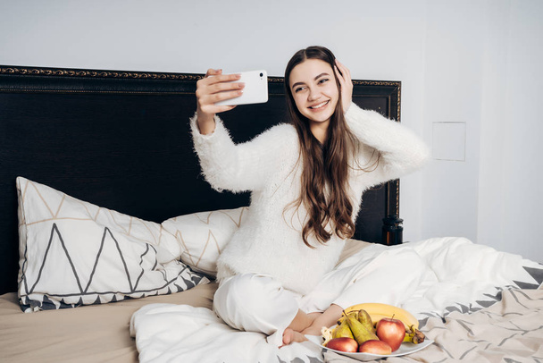 beautiful young girl in white pajamas sits on a bed early in the morning, makes a village and smiles, breakfasts fruit - Foto, immagini
