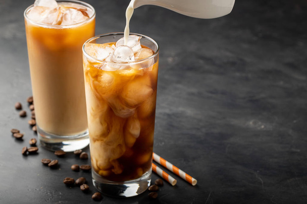 Ice coffee in a tall glass with cream poured over, brown sugar and coffee beans. Cold summer drink on a dark background. With copy space. - Foto, imagen