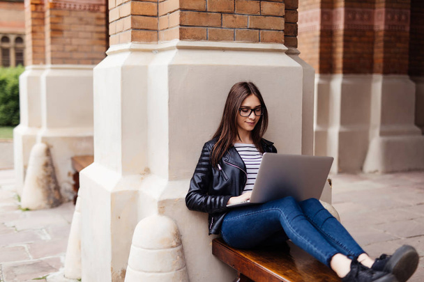 Chica estudiante sonriente sentada en el banco en el parque, trabajando con la computadora portátil y tomando notas, preparándose para los exámenes al aire libre, descansando en el campus universitario, espacio para copiar
 - Foto, imagen