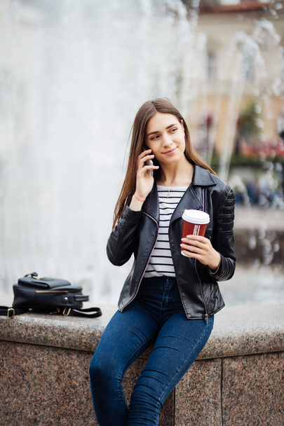 Νεαρό κορίτσι μιλάει στο κινητό με φλιτζάνι καφέ ύπαιθρο κοντά σε σιντριβάνι - Φωτογραφία, εικόνα
