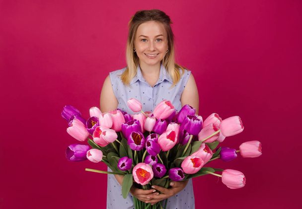 Smiling woman with bunch of flowers in her hands in blue dress. Beautiful woman with bouquet of pink and purple tulips. Summer and spring concept. Isolated on pink background - Photo, image