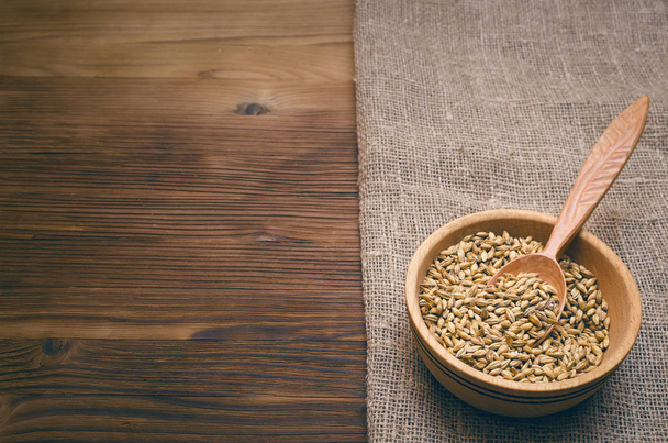 Malt grains in wooden bowl with spoon on burlap cloth on brown wooden table background with copy space. - Fotó, kép