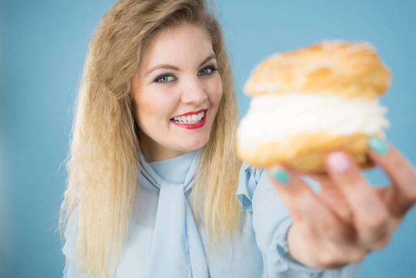 Lekker eten en geluk concept. Grappige vrolijke blonde vrouw met heerlijke choux bladerdeeg taart met slagroom, opgewonden gezicht uitdrukking. Op blauw - Foto, afbeelding