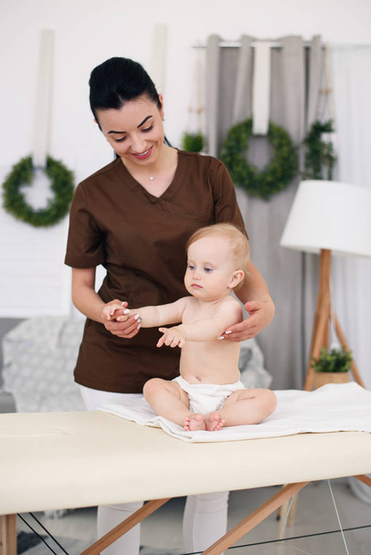 A professional female masseuse makes a massage to a little baby. Children massage on the couch in a modern cozy room. - Фото, изображение