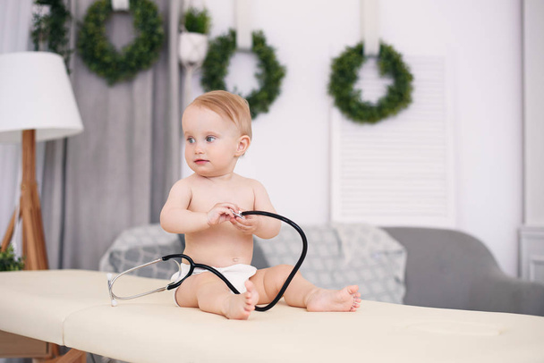 Funny little baby sitting on massage table and playing with stethoscope in modern medical office. - Foto, Imagen