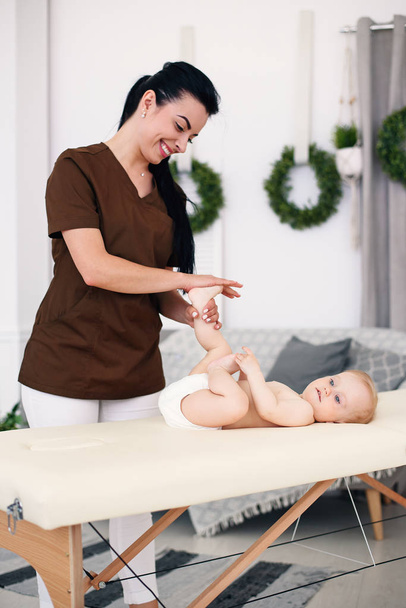 Health care and medical concept. A professional female masseuse makes a massage and gymnastics to a little baby at modern cozy room. - Fotó, kép