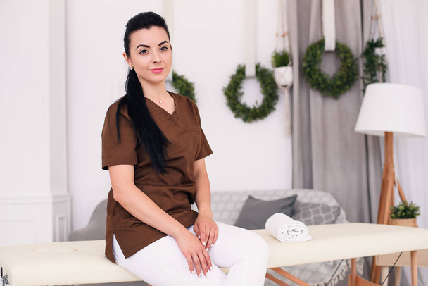 Smiling female doctor looking at camera and sitting on massage table in modern medical office. Professional female masseuse in light room. - Фото, зображення