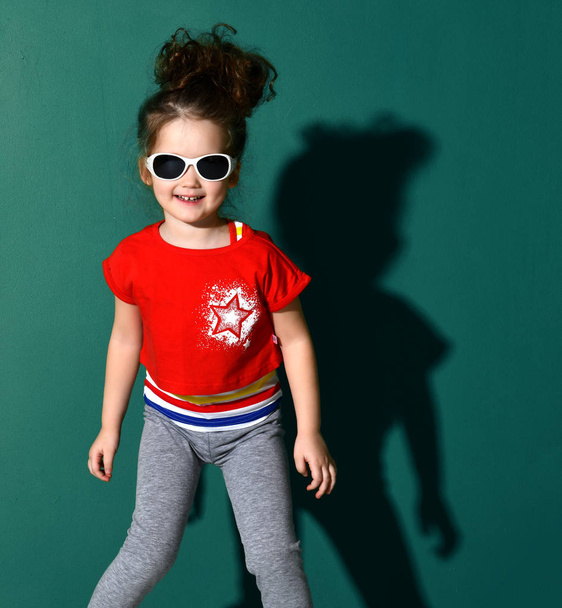 Cute curly baby girl kid in red t-shirt blue jeans and white sunglasses smiling on blue mint  - Foto, imagen
