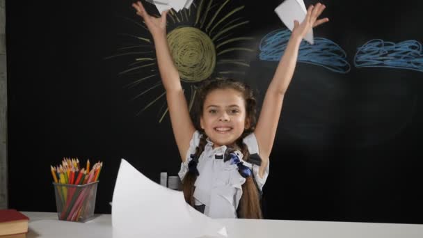 Iskola koncepciója. A vonzó lány ül az asztalon egy táblára a háta mögött. Boldog iskolás dobja papír és mosolyog. Evés - Felvétel, videó