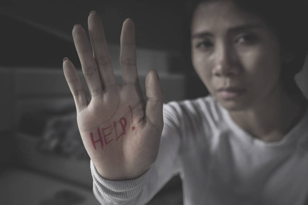 Abused woman looks sad while showing help word on her palm and sitting in the dark room - Foto, afbeelding