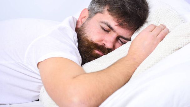 Man in shirt laying on bed, white wall on background. Macho with beard and mustache sleeping, relaxing, having nap, rest. Nap and siesta concept. Guy on calm face sleeping on white sheets and pillow - Foto, afbeelding