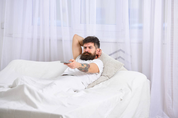 Man laying on bed, watching tv, white curtains on background. Guy on serious face using remote control for switch channel. Macho with beard holds smartphone as controller. Remote control concept - Photo, image