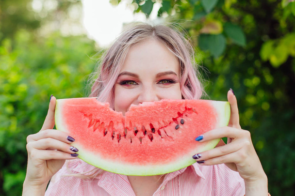 Beautiful young woman with pink hair holding sweet juicy watermelon close to the face like a smile - Foto, Imagen