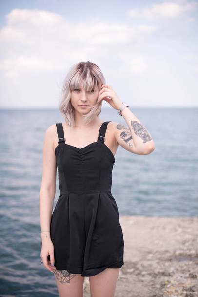 Beautiful young woman with tattoo and black dress posing on sea background - Foto, imagen