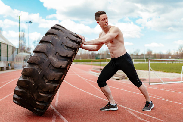 Young muscular man lifting up huge tire for training muscles on stadium - Zdjęcie, obraz