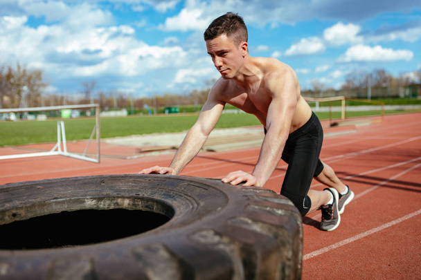 Young muscular man lifting up huge tire for training muscles on stadium - Photo, Image
