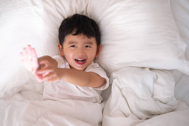 3 years old little cute Asian boy in white shirt at home on the bed, kid lying playing and smiling on white bed with pillow and blanket, top view with copy space for add text. - Fotografie, Obrázek