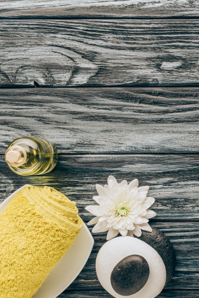 flat lay with spa and massage treatment arrangement with towel, essential oil, pebbles and chrysanthemum flower on wooden background - Photo, Image