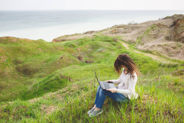 Молодая деловая женщина или студентка в повседневной одежде с трепещущими волосами, сидящая на траве с помощью ноутбука в поле, раздвигает руки, работая на свежем воздухе на зеленом фоне. Мобильный офис. Концепция образа жизни
 - Фото, изображение