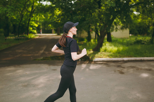 Side view Young athletic brunette girl in black uniform and cap training, doing sport exercises and running, looking straight on path in city park outdoors. Fitness, healthy lifestyle concept - Photo, Image