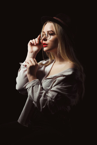 Tender blonde woman in glasses and hat, wearing shirt with naked shoulders, posing with dramatic light at studio - Photo, Image