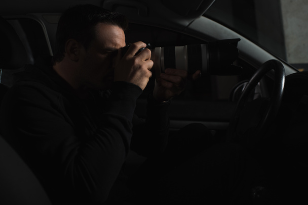side view of concentrated man doing surveillance by camera with object glass from his car  - Photo, Image