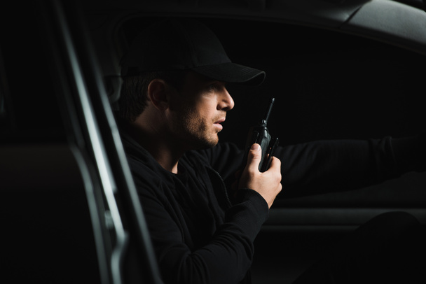 side view of male paparazzi in cap doing surveillance and using talkie walkie in his car - Fotoğraf, Görsel