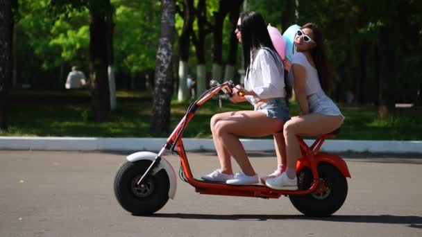 Two young and sexy brunette friends with loose hair in short denim shorts riding an electric motorcycle in the Park on a Sunny day enjoying hugging each other. Best friends spend time together - Imágenes, Vídeo