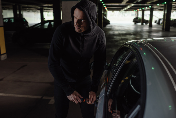 cautious male robber in black hoodie intruding car by screwdriver  - 写真・画像