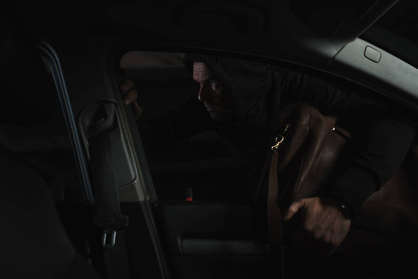 προσεκτική αρσενικό κλέφτης στο μαύρο hoodie κλέβοντας την τσάντα από το αυτοκίνητο - Φωτογραφία, εικόνα