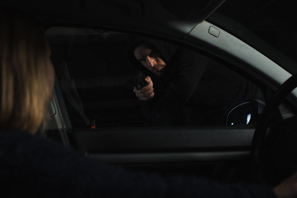 αρσενικό ληστής σε μαύρο hoodie με στόχο με όπλο την γυναίκα να κάθεται στο αυτοκίνητο - Φωτογραφία, εικόνα