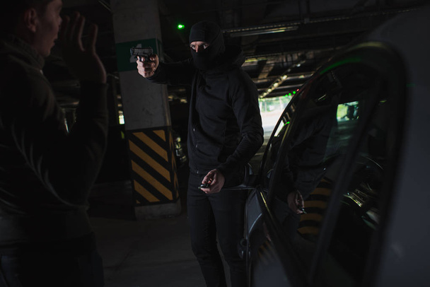 male mugger in balaclava aiming by gun on man standing near his car - 写真・画像
