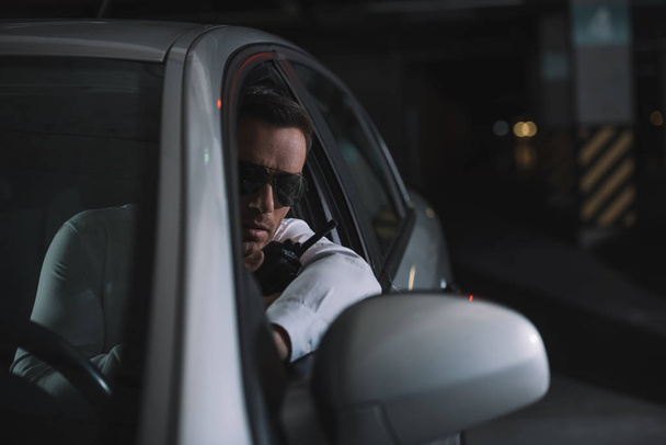 σοβαρή ανδρική μυστικός πράκτορας σε γυαλιά ηλίου κάνει επιτήρηση και γουόκι τόκι που χρησιμοποιεί στο αυτοκίνητο - Φωτογραφία, εικόνα