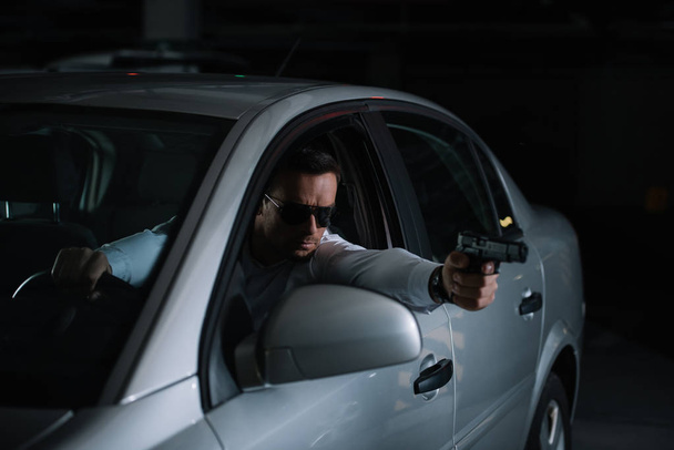 male private detective in sunglasses aiming by gun from car - Fotó, kép